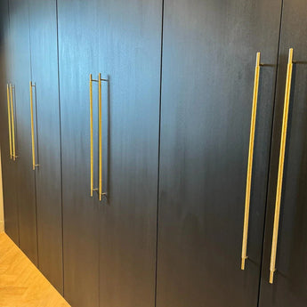 Une rangée d'armoires fermées Remo Gold Deluxe de yessential.fr, avec du bois foncé et de longues poignées dorées, se dresse contre un parquet en bois clair.