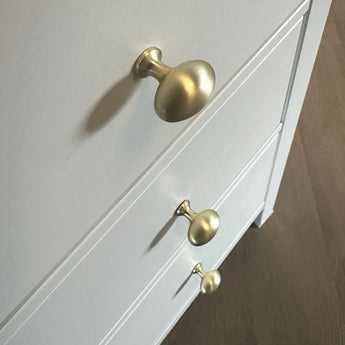 Gros plan de trois poignées Agero Gold Knob Deluxe de yessential.fr sur une armoire blanche, placée en diagonale au-dessus d'un plancher en bois marron.