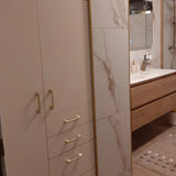 Créez une salle de bain moderne avec les carreaux effet marbre Ramiro Gold Deluxe de yessential.fr, une armoire haute avec des poignées dorées, un meuble sous-vasque en bois et un lavabo blanc, un miroir au-dessus et un tapis à motifs pour une finition élégante.