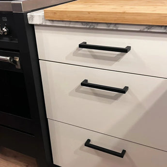 Un meuble de cuisine avec trois tiroirs blancs, chacun équipé d'une poignée Vasco Black de yessential.fr. Le plan de travail allie marbre et bois et une partie d'un four noir est visible à gauche.