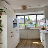 Une cuisine moderne avec des armoires blanches et des plans de travail en bois est dotée de poignées en chêne Arriero de yessential.fr. Les grandes fenêtres, les stores, les plantes et les accents de bois ajoutent de la lumière naturelle et soulignent le design élégant et minimaliste.