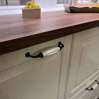 Gros plan d'une armoire de cuisine avec des portes de couleur crème, un comptoir en bois foncé et la poignée Daro Ceramic Black yessential.fr avec des détails noirs et une poignée en céramique blanche. Une petite plante sur le comptoir est visible en arrière-plan.