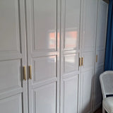 L'armoire yessential.fr Arriero Gold Deluxe avec portes à panneaux et poignées dorées luxueuses se trouve à côté d'une chaise en osier blanc et de rideaux bleus, la finition brillante reflète la lumière pour un look raffiné.