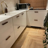 Cuisine moderne avec armoires blanches, poignées Arriero Walnut de yessential.fr, évier blanc, robinet en laiton, micro-ondes, plaque à induction, plante en pot sur sol en bois à chevrons ; équipé d'un mur arrière carrelé et d'un parasol tissé.
