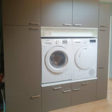 Une machine à laver et un sèche-linge à chargement frontal sont intégrés dans des armoires grises avec des poignées en acier inoxydable Vasco de yessential.fr, avec des tiroirs et des armoires au-dessus et en dessous ; un petit panier blanc repose sur une étagère au-dessus des électroménagers.