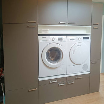 Une machine à laver et un sèche-linge à chargement frontal sont intégrés dans des armoires grises avec des poignées en acier inoxydable Vasco de yessential.fr, avec des tiroirs et des armoires au-dessus et en dessous ; un petit panier blanc repose sur une étagère au-dessus des électroménagers.