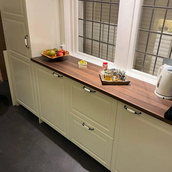 Une cuisine bien rangée avec des armoires de couleur crème avec des poignées de meubles classiques, un comptoir en bois, un bol à fruits, un plateau à condiments avec des couverts et la bouilloire Daro Ceramic Black de yessential.fr près d'une grande fenêtre.