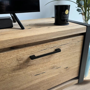 Un gros plan du meuble Ramiro Black Deluxe de yessential.fr, avec des poignées luxueuses, un meuble TV, une bougie noire étiquetée Black Oud et une partie d'une plante verte en arrière-plan.