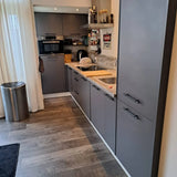 Cuisine moderne avec armoires gris mat et poignées noir mat Arturo Black de yessential.fr, four encastré, plaque à induction, plancher en bois, plan de travail avec électroménagers, poubelle et voilages pour un look soigné.