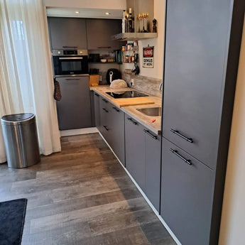 Cuisine moderne avec armoires gris mat et poignées noir mat Arturo Black de yessential.fr, four encastré, plaque à induction, plancher en bois, plan de travail avec électroménagers, poubelle et voilages pour un look soigné.