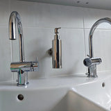 Le lavabo double Alberto Chrome de yessential.fr est doté de deux robinets chromés et d'un élégant distributeur de savon, montés au mur au-dessus d'un comptoir blanc contre un mur arrière carrelé.