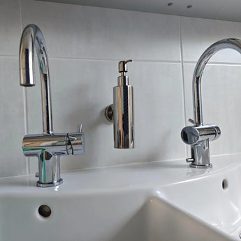 Le lavabo double Alberto Chrome de yessential.fr est doté de deux robinets chromés et d'un élégant distributeur de savon, montés au mur au-dessus d'un comptoir blanc contre un mur arrière carrelé.