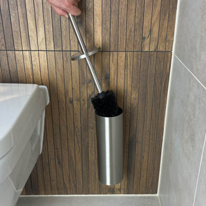 Une main tient la brosse supplémentaire yessential.fr à tête remplaçable au-dessus d'un support en acier inoxydable sur un mur lambrissé, à côté de toilettes avec un siège blanc et un sol carrelé - prêtes à être nettoyées.