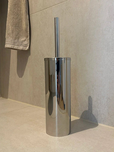La brosse WC avec support Bruno Chrome de yessential.fr est posée sur un sol carrelé beige contre le mur, avec une serviette beige sur le mur en arrière-plan.
