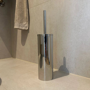 La brosse WC avec support Bruno Chrome de yessential.fr est posée sur un sol carrelé beige contre le mur, avec une serviette beige sur le mur en arrière-plan.