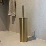 L'ensemble de toilettes Tygo Gold (sans perçage) de yessential.fr - un porte-brosse de toilette de couleur dorée avec poignée assortie - repose sur du carrelage clair avec une serviette grise et est facile à installer sans perçage, parfait pour vos accessoires de salle de bain.