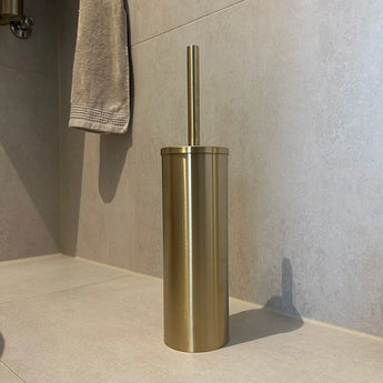 L'ensemble de toilettes Juano Gold (sans perçage) de yessential.fr, avec une brosse de toilette dorée et un support cylindrique, repose sur un carrelage de salle de bain clair avec une serviette grise en arrière-plan.