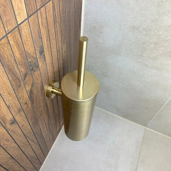Le porte-brosse de toilette Branco Gold de yessential.fr est monté sur un mur avec du carrelage imitation bois marron, à côté du carrelage gris clair au sol et au mur dans un coin de salle de bain.