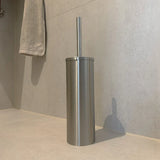 L'ensemble de toilettes Ludo en acier inoxydable (sans perçage) de yessential.fr, doté d'un porte-brosse de toilette en acier inoxydable avec poignée assortie, est placé sur le sol de la salle de bain légèrement carrelé, à proximité d'une serviette grise accrochée au mur.