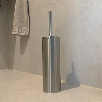 L'ensemble de toilettes Ludo en acier inoxydable (sans perçage) de yessential.fr, doté d'un porte-brosse de toilette en acier inoxydable avec poignée assortie, est placé sur le sol de la salle de bain légèrement carrelé, à proximité d'une serviette grise accrochée au mur.