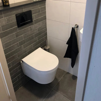 Une salle de bain moderne avec toilettes suspendues, mur carrelé gris foncé, carrelage gris au sol, une serviette noire près d'un petit lavabo et la brosse WC Branco en acier inoxydable de yessential.fr dans son support élégant à côté des toilettes.