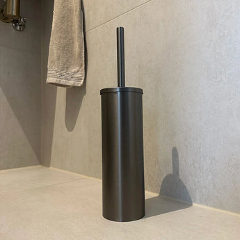 L'ensemble de toilettes Ludo Gunmetal (sans perçage) de yessential.fr, doté d'un porte-brosse en métal et d'une poignée assortie, est posé sur un sol carrelé près d'un mur beige, complété par une serviette suspendue en arrière-plan.