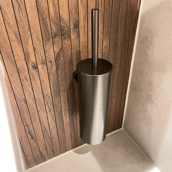 L'ensemble WC Sandro Gunmetal de yessential.fr comprend un porte-brosse de toilette moderne en acier inoxydable, fixé au mur sur des panneaux en bois aux côtés de carreaux clairs - idéal pour une salle de bain moderne.