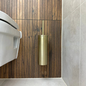 Des toilettes blanches se trouvent près d’un mur avec des panneaux de bois verticaux. Le porte-brosse de toilette yessential.fr Branco Gold est monté au mur, et le sol et le mur à côté ont des carreaux gris clair.