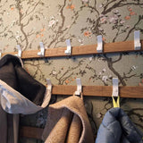 Le 10x Huso White de yessential.fr, un porte-manteau moderne en bois avec crochets blancs, est monté sur un papier peint à motif floral et offre de la place pour plusieurs manteaux soigneusement accrochés dans des couleurs neutres.