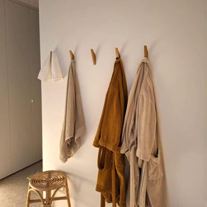 Trois peignoirs et une serviette sont accrochés au porte-manteau Fabio Oak avec crochets de yessential.fr contre un mur blanc. A proximité se trouve un tabouret en bois avec un siège tressé, tandis qu'une lumière chaude sur la gauche crée une atmosphère chaleureuse dans la pièce.