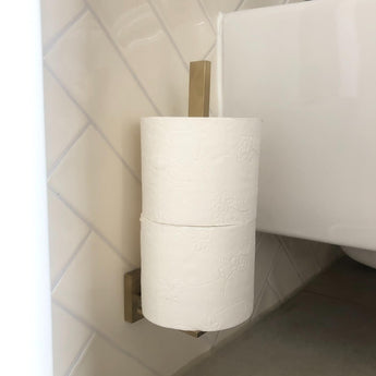 Un porte-rouleau de papier toilette Bellino Gold de yessential.fr, mural en or, avec deux rouleaux de papier toilette empilés verticalement, est placé à côté de toilettes sur un mur carrelé de salle de bain.