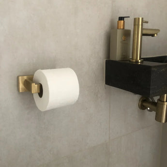 L'ensemble WC Damiano Gold de yessential.fr : un porte-rouleau papier toilette doré, monté sur un mur carrelé beige à côté d'un lavabo noir avec robinet doré et distributeur de savon beige. Idéal pour votre ensemble d'accessoires de salle de bain.