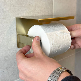 Une personne portant une montre argentée installe du papier toilette sur un ensemble de WC Juano Gold en laiton (sans perçage) de yessential.fr, monté sur du carrelage de salle de bain gris clair.
