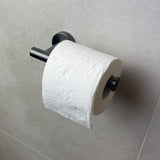 Un rouleau de papier toilette avec un motif en relief ondulé est suspendu au porte-rouleau de papier toilette Tino Gunmetal de yessential.fr, monté sur un mur carrelé gris clair pour un ajout élégant et moderne à vos accessoires de salle de bain.
