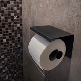 Un rouleau de papier toilette se trouve sur le porte-rouleau de papier toilette Tazio Black de yessential.fr, monté sur un mur de salle de bains carrelé gris avec des carreaux de mosaïque visibles à proximité.