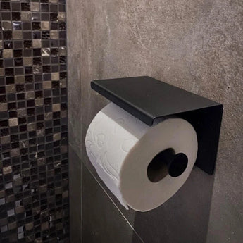 Un rouleau de papier toilette se trouve sur le porte-rouleau de papier toilette Tazio Black de yessential.fr, monté sur un mur de salle de bains carrelé gris avec des carreaux de mosaïque visibles à proximité.