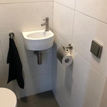 Un petit lavabo blanc avec un robinet argenté se dresse dans un coin carrelé au-dessus d'un sol gris. Une serviette noire est suspendue à proximité et un porte-rouleau de papier toilette en acier inoxydable yessential.fr Julio est monté près de l'évier. Un bouton carré affleurant est accroché au mur.