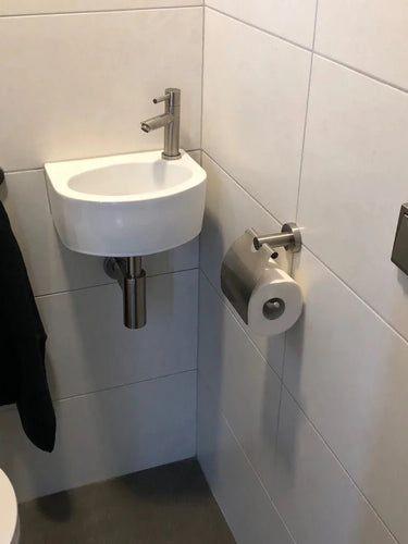 Un petit lavabo blanc avec un robinet argenté se dresse dans un coin carrelé au-dessus d'un sol gris. Une serviette noire est suspendue à proximité et un porte-rouleau de papier toilette en acier inoxydable yessential.fr Julio est monté près de l'évier. Un bouton carré affleurant est accroché au mur.