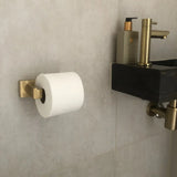 Le porte-rouleau de papier toilette Vero Gold Deluxe de yessential.fr est monté sur un mur carrelé à côté d'un lavabo noir moderne avec des luminaires en laiton et un distributeur de savon, apportant des accessoires de salle de bain de luxe dans l'espace.