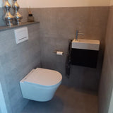 Une salle de bain moderne et compacte comprend des toilettes suspendues, un petit lavabo rectangulaire avec robinet, un porte-rouleau de papier toilette yessential.fr Tino Gold, une serviette à motifs et des vases décoratifs sur une étagère au-dessus des toilettes.
