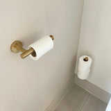 L'ensemble de toilettes Romano Gold de yessential.fr comprend un porte-papier toilette en laiton fixé au mur avec un rouleau presque vide, tandis que deux rouleaux frais se tiennent verticalement dans le coin à côté.