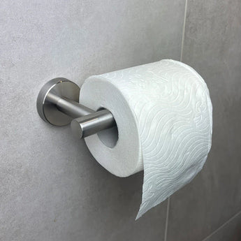 Un rouleau de papier toilette est suspendu à un porte-rouleau de papier toilette Tino en acier inoxydable de yessential.fr, monté sur un mur carrelé gris clair. Le motif en papier gaufré et le support moderne ajoutent du style parmi d'autres accessoires de toilette élégants.