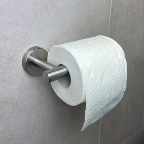 Un rouleau de papier toilette est suspendu à un porte-rouleau de papier toilette Tino en acier inoxydable de yessential.fr, monté sur un mur carrelé gris clair. Le motif en papier gaufré et le support moderne ajoutent du style parmi d'autres accessoires de toilette élégants.