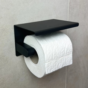 Un rouleau de papier toilette blanc avec un subtil motif en relief est monté sur le porte-papier toilette Tazio Black (No Drill) yessential.fr contre un mur de salle de bains carrelé gris clair.