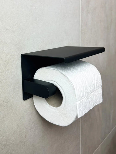 Un rouleau de papier toilette blanc gaufré sur un porte-rouleau de papier toilette yessential.fr Tazio Black avec étagère, fixé à un mur carrelé de salle de bains.