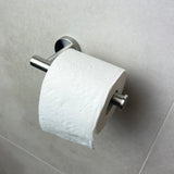 Un rouleau de papier toilette avec un motif en relief ondulé et floral repose sur le Tino Stainless Steel de yessential.fr, monté sur un mur carrelé gris clair. Ce support élégant donne à votre collection d'accessoires de toilette un look élégant.