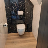 Une salle de bain moderne avec les toilettes murales yessential.fr Tino Black, paroi arrière en marbre noir, murs blancs, parquet en bois, accessoires noir mat et un porte-rouleau papier toilette en acier inoxydable sur le mur de gauche.