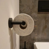 Un rouleau de papier toilette avec des taches bleues est suspendu à l'ensemble de toilettes Cosimo Black de yessential.fr, monté sur un mur blanc avec un mur texturé gris et un ensemble d'accessoires de salle de bain en arrière-plan.