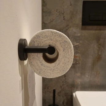 Un rouleau de papier toilette avec des taches bleues est suspendu à l'ensemble de toilettes Cosimo Black de yessential.fr, monté sur un mur blanc avec un mur texturé gris et un ensemble d'accessoires de salle de bain en arrière-plan.
