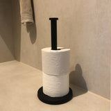 Un porte-rouleau de papier toilette Jurado Black de yessential.fr se dresse verticalement dans le coin d'une salle de bain carrelée beige, contenant deux rouleaux de papier toilette blanc, avec une serviette soigneusement pliée sur le mur au-dessus.